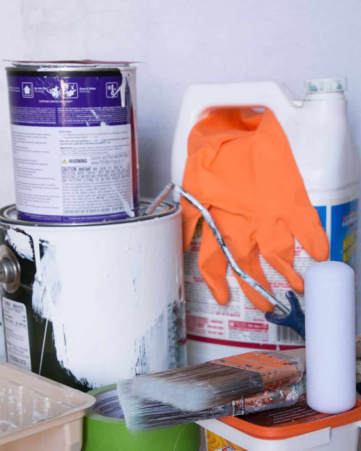 cabinet painting supplies, can of primer, paint, orange rubber glove, small foam roller, wood filler and painter's tape.