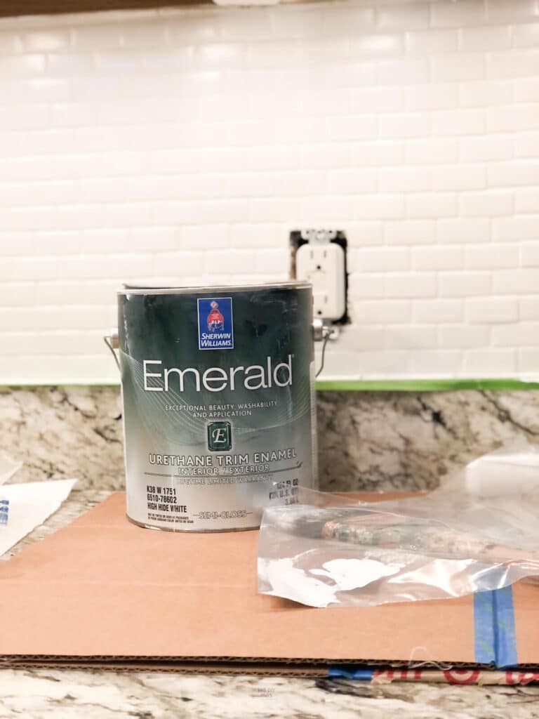 gallon of Emerald Trim Enamel paint on cardboard in kitchen with brush in front of white backsplash and cabinets.