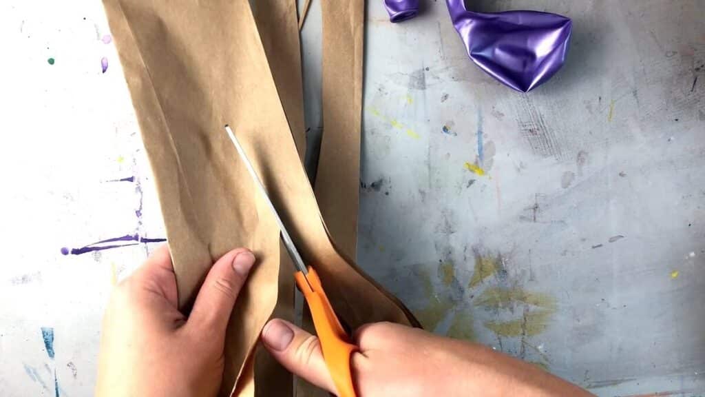 hands holding orange scissors cutting brown paper bags into strips.