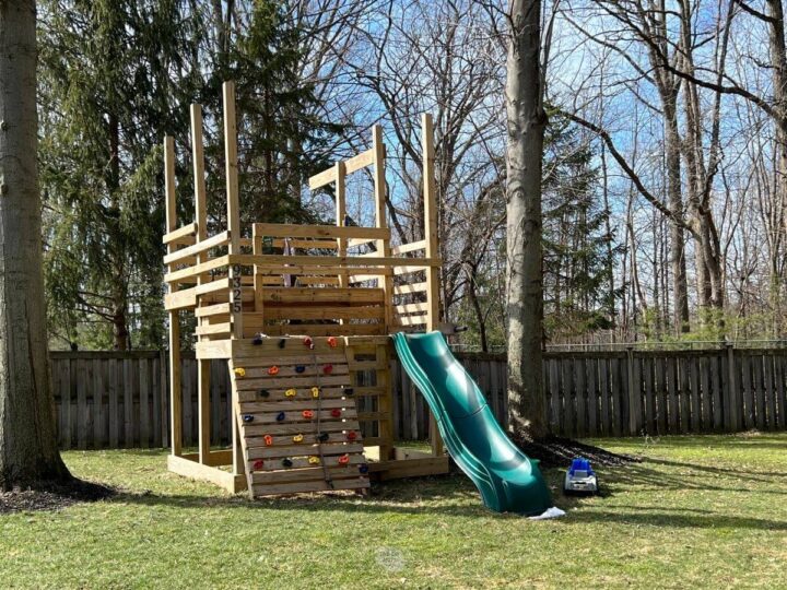 wooded DIY playset with green slide, rockwall and two platform levels in backyard.