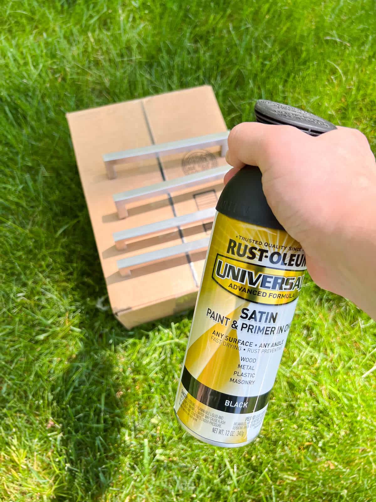 Hand holding black satin Rustoleum spray paint with kitchen hardware on grass.
