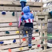 boy in superhero sweatshirt climbing wooden rock wall with colorful rock wall holds.