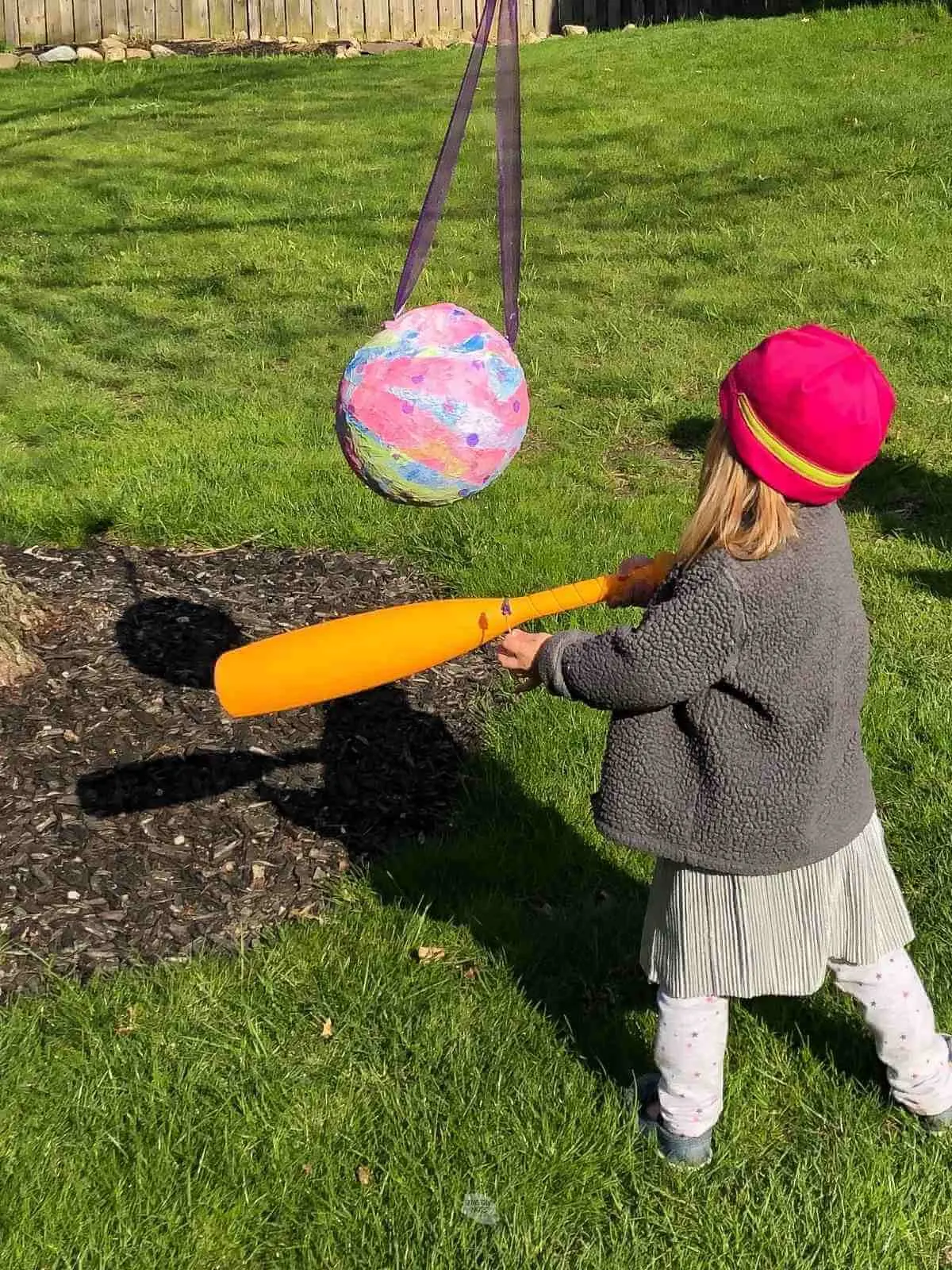 little girl in pink hat and gray coat swinging plastic bat at homemade piñata.