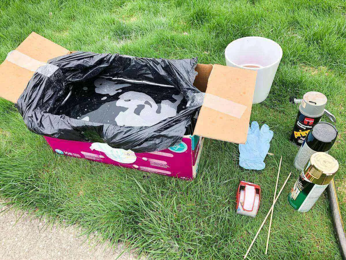diaper box with bag filled with water, flower pot, spray paint, hose and packing tape on grass.