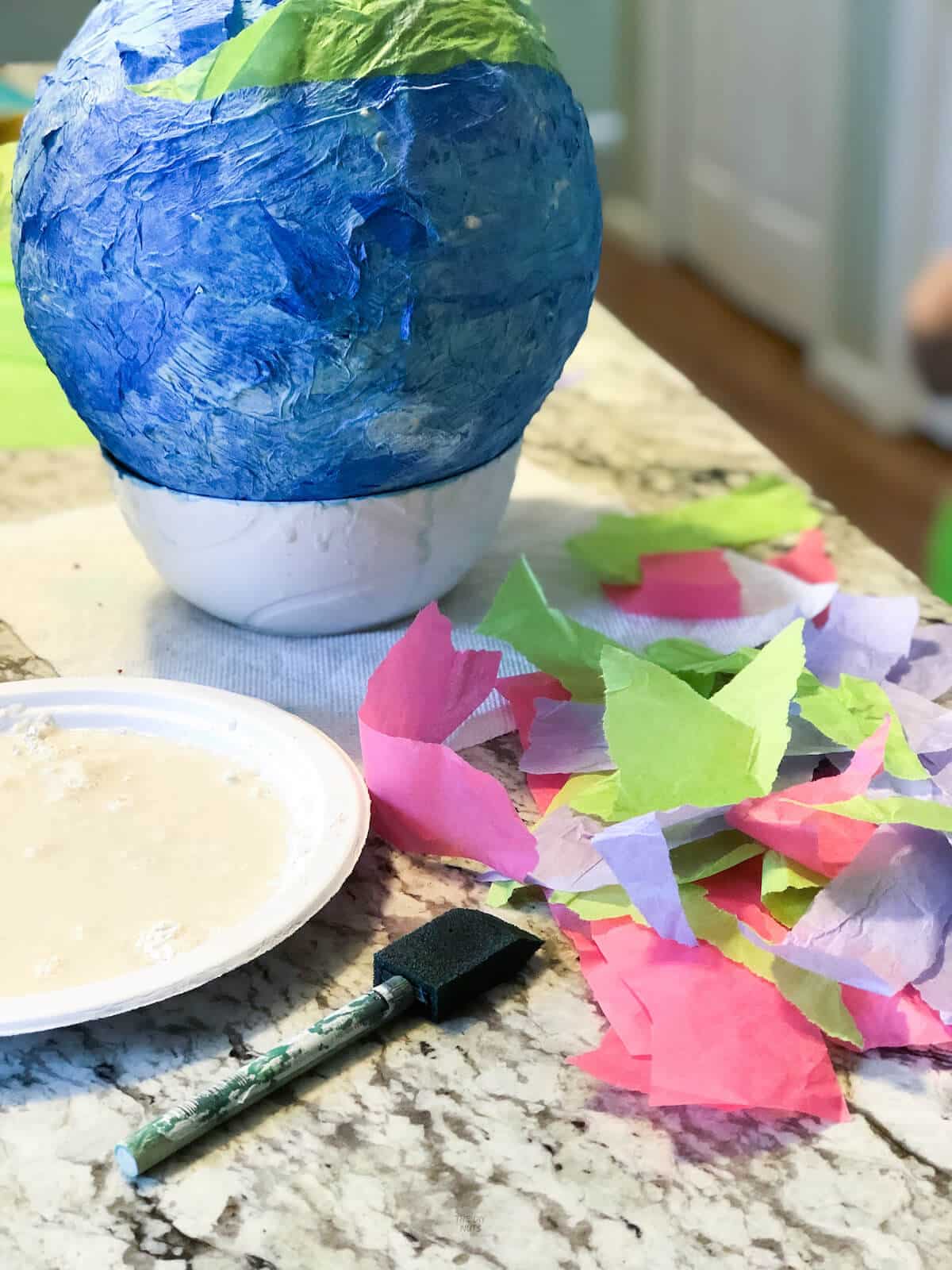 blue homemade piñata in bowl with plate of paste and loose tissue paper on counter.