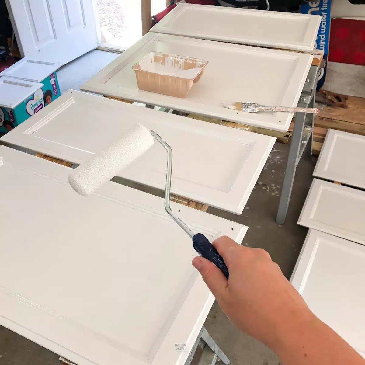 hand holding small roller with cabinet doors on sawhorses and white paint and brush.