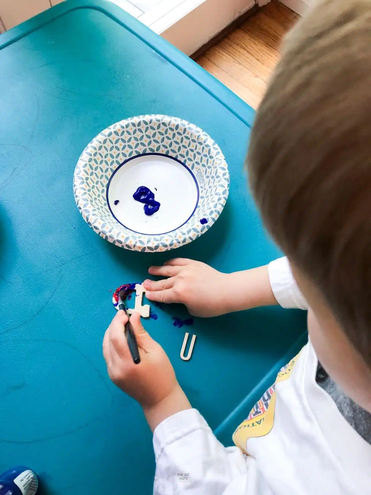 little boy painting small wooden letters with blue paint on teal table.