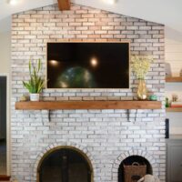 Final white washed large brick fireplace with wood beam and shelves.