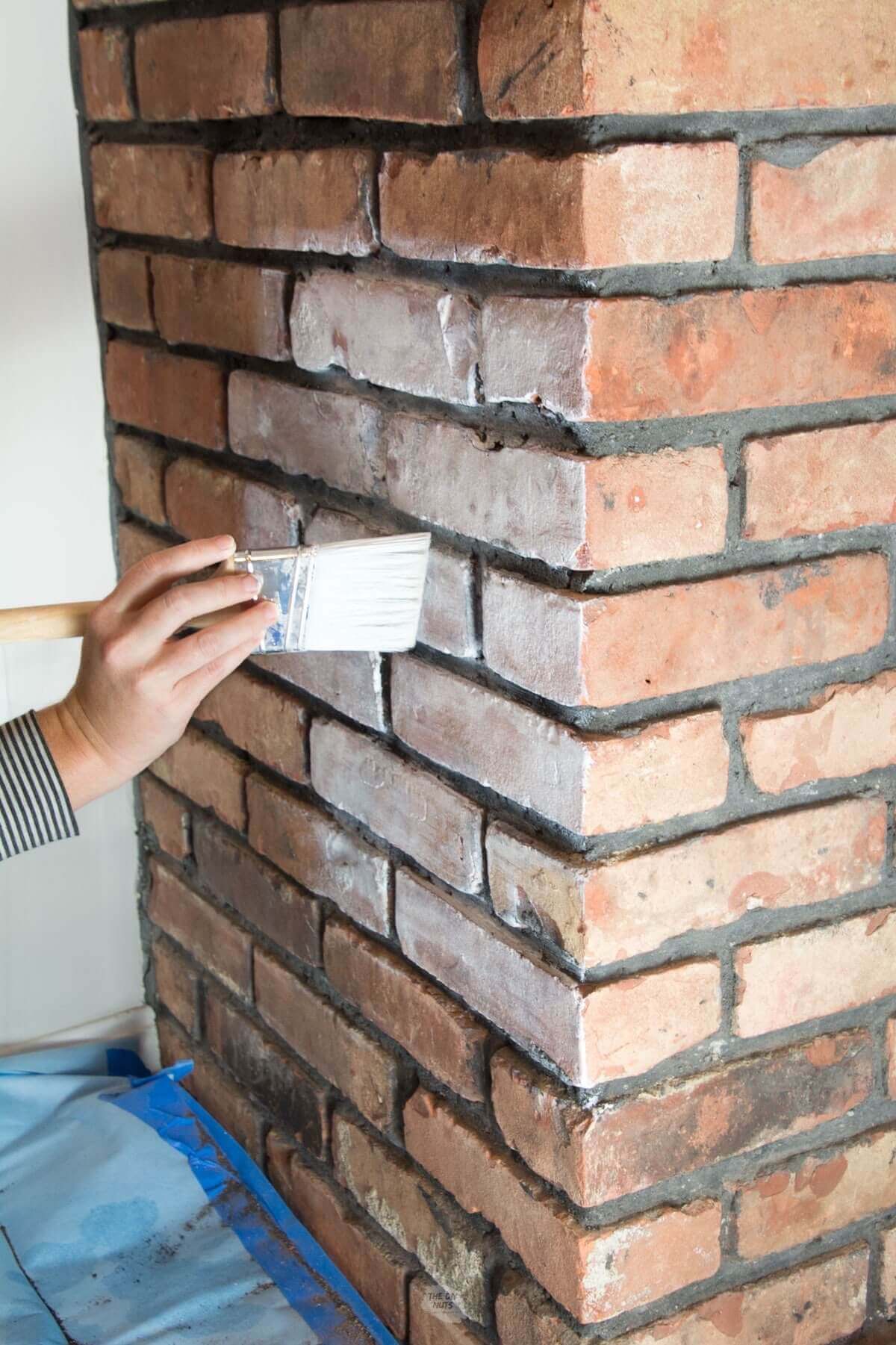 hand holding paint brush and putting white paint on brick fireplace.