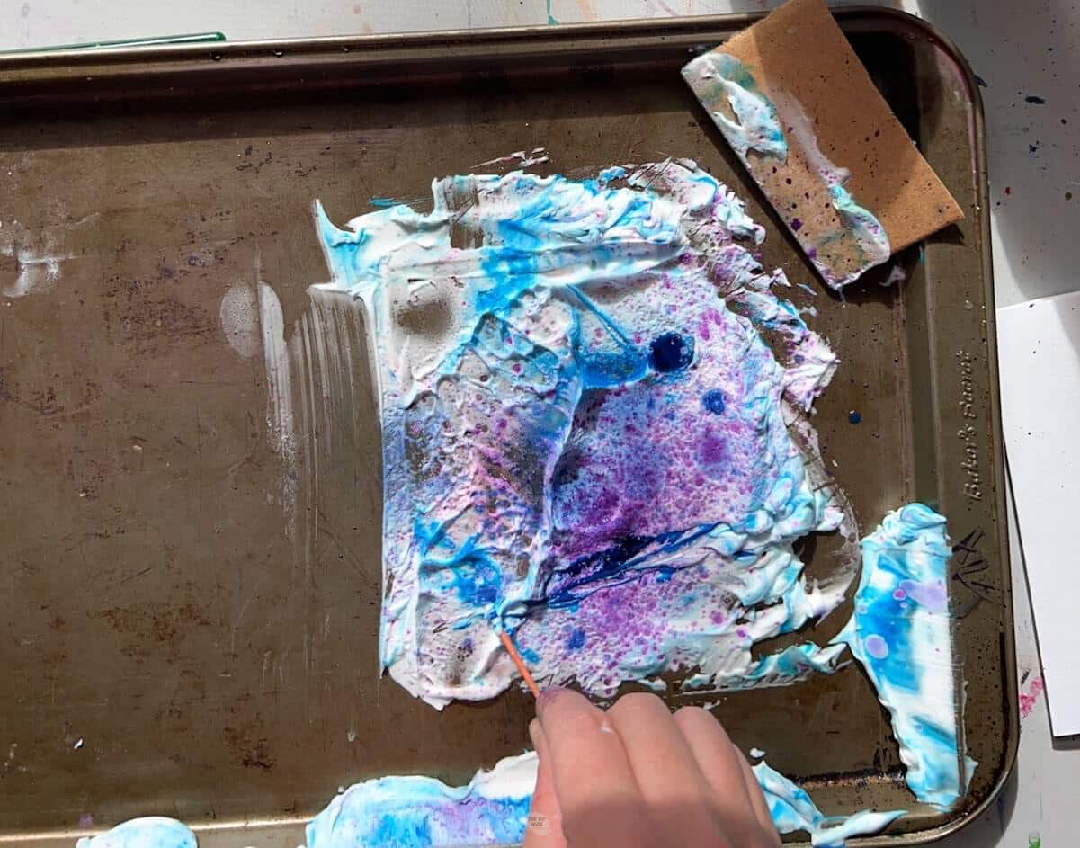 hand using toothpick to swirl blue and purple paint in shaving cream.
