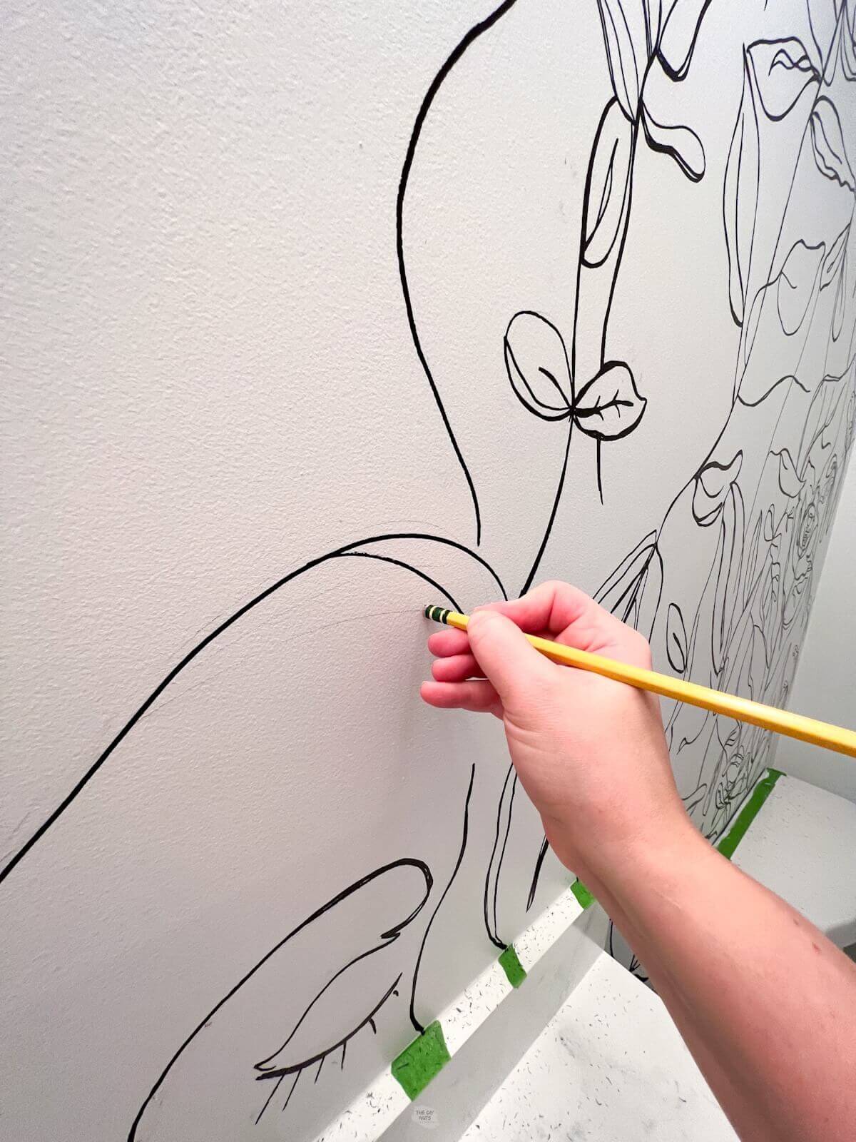 hand holding yellow pencil using eraser to erase pencil lines on white wall with black marker drawing on it.