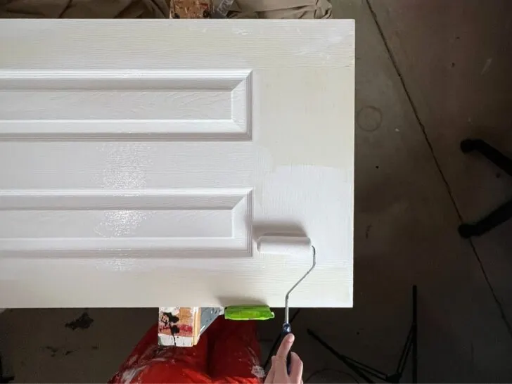 hand holding small roller adding white paint interior door on sawhorses.